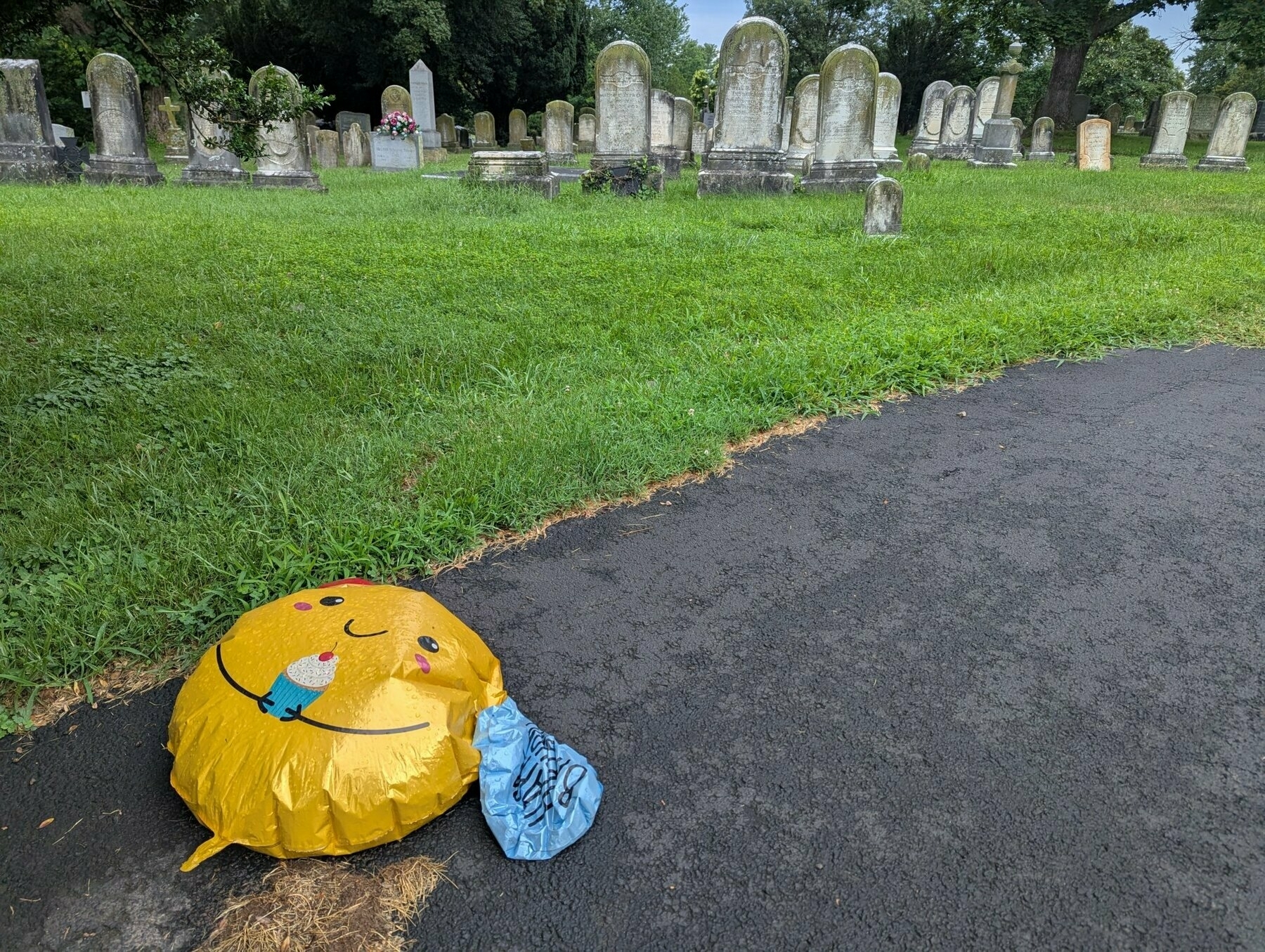 Deflated happy birthday balloon with a smiley face and cupcake, laying on the ground in a cemetery