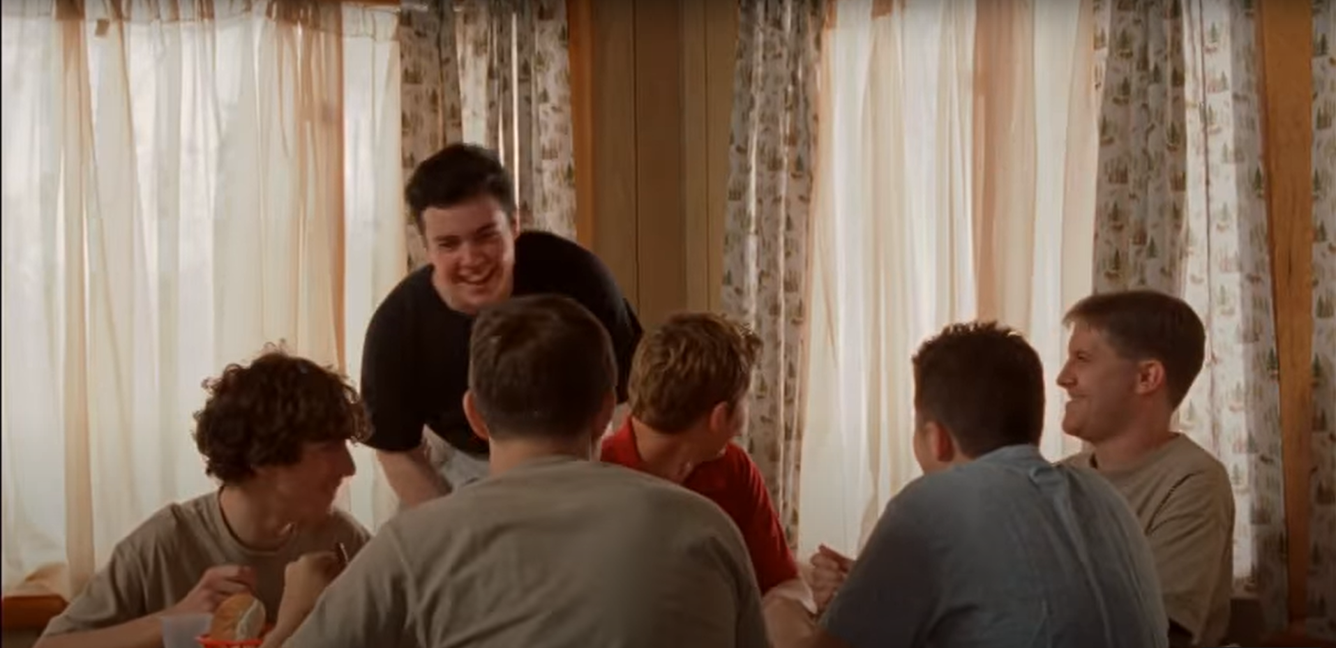 A group of six people sit around a table, with one person standing and smiling, in a room with patterned curtains. I am laughing.