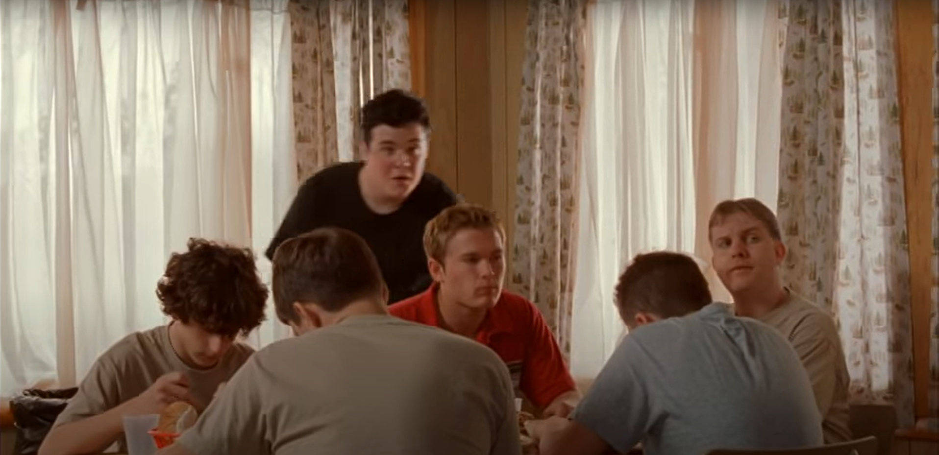 A group of six people sit around a table, with one person standing and smiling, in a room with patterned curtains. I am looking inquisitive.